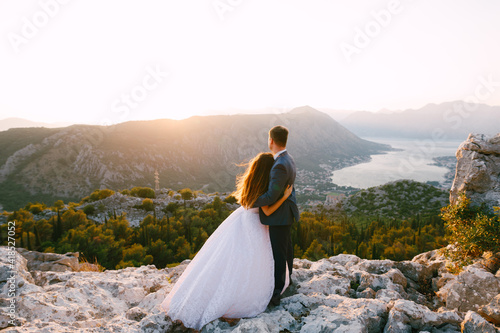 A bride and a groom stand hugging on the mount Lovcen and look at the panoramic view of the Bay of Kotor