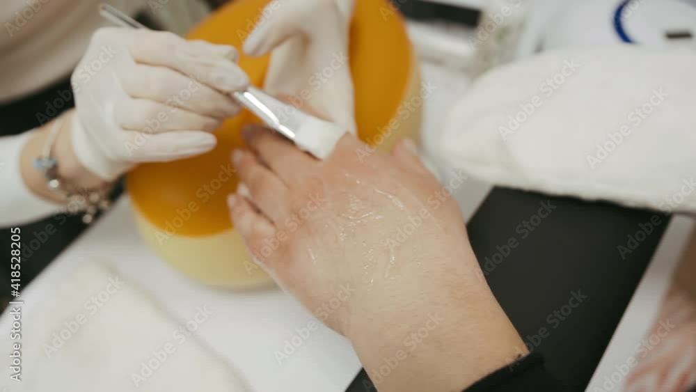 A beautician performs a paraffin therapy procedure and uses a brush apply mask to the client's hand and gently smears over the entire area from dryness and age-related skin changes. A beautician performs a paraffin therapy procedure and uses a brush apply mask to the client's hand and gently smears over the entire area from dryness and age-related skin changes.