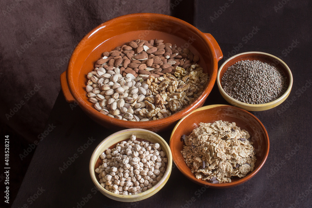 A large artisan clay pot with almonds, pistachios, and walnuts along with three bowls filled with lentils, chickpeas, and toasted flakes of rice, whole wheat, and chocolate on a wooden table. A large artisan clay pot with almonds, pistachios, and walnuts along with three bowls filled with lentils, chickpeas, and toasted flakes of rice, whole wheat, and chocolate on a wooden table.