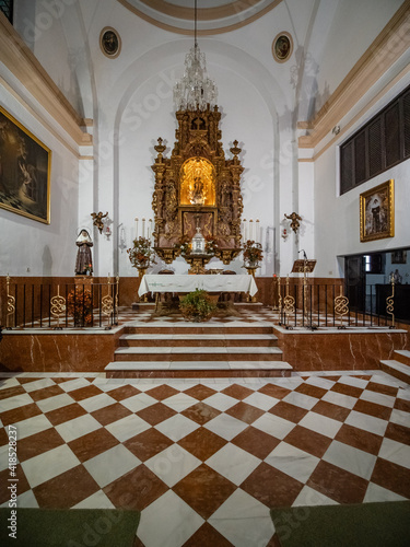 Photography Interior view of a side chapel in the Church of Santa Maria la Mayor in Ronda, Spain, showing a gilded Baroque altarpiece, marble steps, and a brown and white checkered tile floor