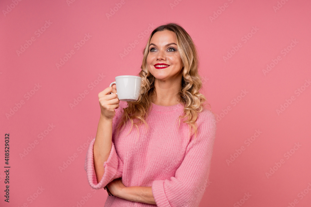 Drink tea, enjoy a hot drink. A young woman in a warm pink sweater on a pink background with a white mug in her hands. Drink tea, enjoy a hot drink. A young woman in a warm pink sweater on a pink background with a white mug in her hands.
