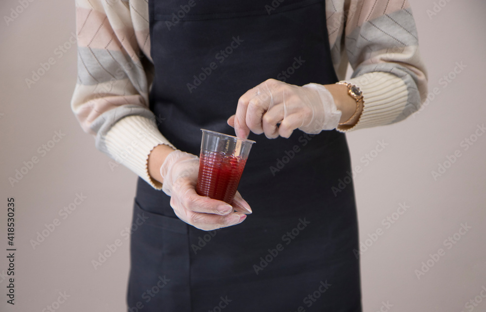 a girl in a gray apron and gloves mixes a red viscous liquid with a wooden stick, in a plastic cup, in a bright apartment, during the day. Master class, experiments, mixing