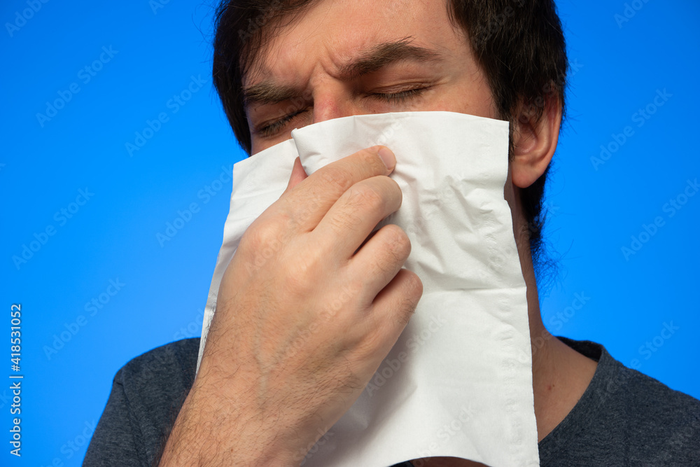 Caucasian male sneezing blowing nose into white paper tissue close up studio shot isolated on blue background Caucasian male sneezing blowing nose into white paper tissue close up studio shot isolated on blue background