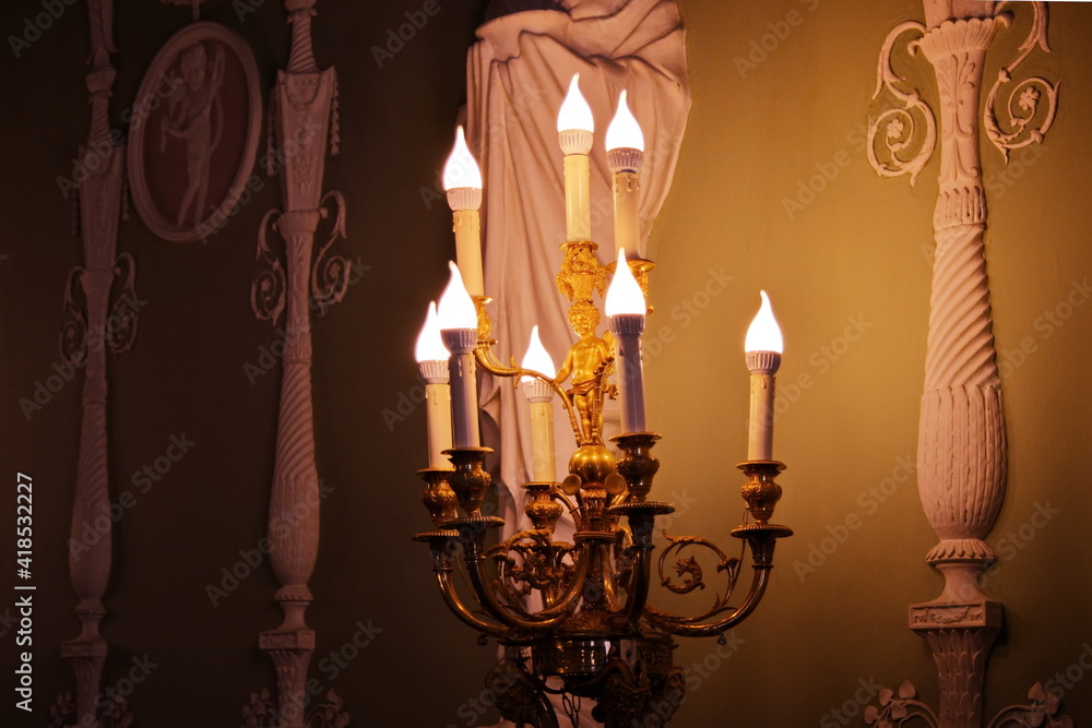 Retro interior detail - classic candelabrum with electric light bulbs in the shape of candles glowing in a dark room with antique design and stucco decorations