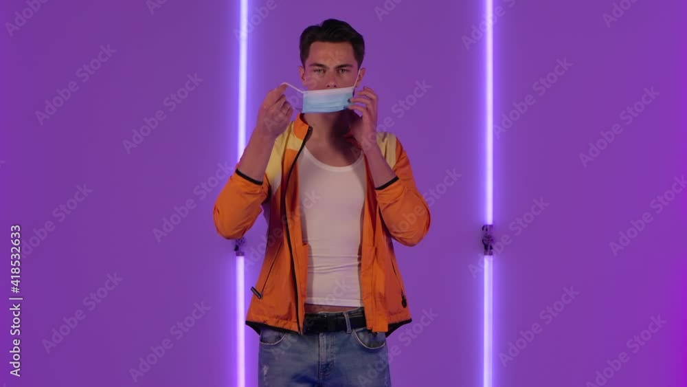 Portrait of stylish guy puts on and then removes the medical protective mask. Male fashion model in yellow jacket poses in dark studio against the backdrop of bright neon lights. Slow motion. Portrait of stylish guy puts on and then removes the medical protective mask. Male fashion model in yellow jacket poses in dark studio against the backdrop of bright neon lights. Slow motion.