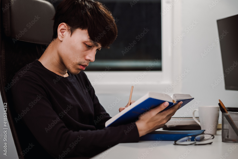 Young handsome asian man reading book and writing note at work desk late at night, Knowledge and learning concept. Young handsome asian man reading book and writing note at work desk late at night, Knowledge and learning concept.
