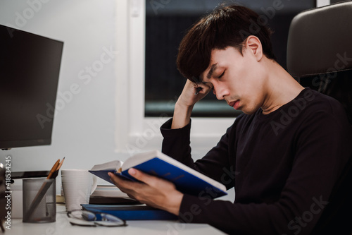 Young handsome asian man sleeping from heavy reading book at work desk late at night, Knowledge and learning concept.