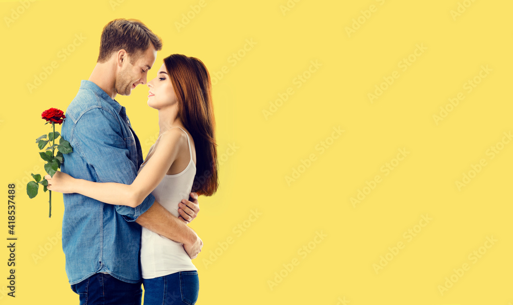 Naklejka premium Love, relationship, dating, flirting, romantic concept - amorous hugging couple with flower, standing close to each other. Studio portrait of young man and woman, isolated over yellow background.