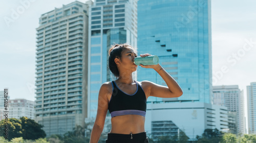 Taking a workout break: Tired woman in in activewear standing and drinking water after exercise with modern buildings in the background. Beautiful young female runner sipping water from blue bottle