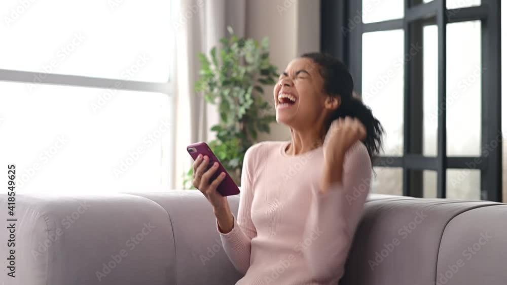 Surprised African American teenage girl sitting on the couch, looking at mobile phone, checking social media messages or browsing the internet and cannot believe her eyes, unbelievable news concept Surprised African American teenage girl sitting on the couch, looking at mobile phone, checking social media messages or browsing the internet and cannot believe her eyes, unbelievable news concept