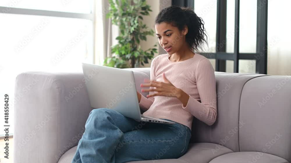 Young mixed-race woman with Afro hair greeting by video call and smiling, sitting on a sofa at home, work from home, conversation with colleagues, business partners, waving hand, video meeting concept Young mixed-race woman with Afro hair greeting by video call and smiling, sitting on a sofa at home, work from home, conversation with colleagues, business partners, waving hand, video meeting concept