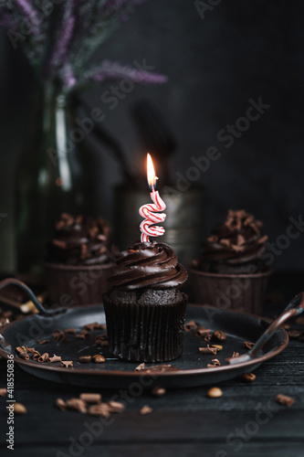 Several muffins or cupcakes with chocolate shaped cream at black table. Festive candle burns on a chocolate cake.