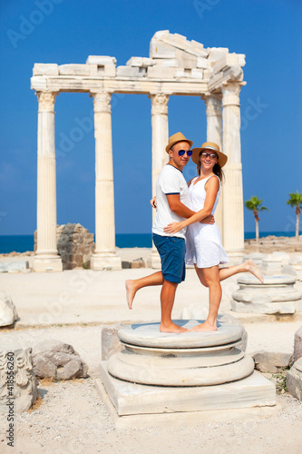 A cheerful and beautiful tanned man and woman in hats and sunglasses embrace at the temple of Apollo in Turkey.