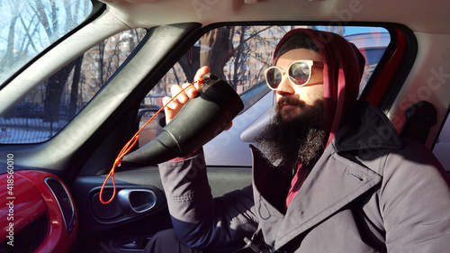 Close up portrait of a bearded young man drinking takeaway coffee. A bearded man with a mustache looks out the window in the car. Man in the car drinking coffee © sarymsakov.com