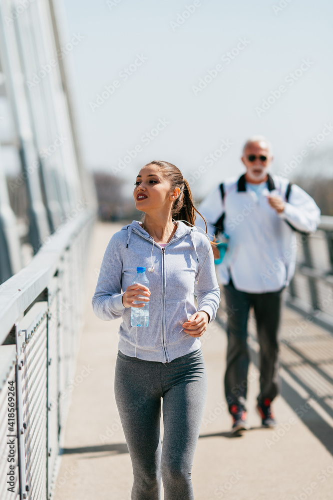 Fit and tired senior man in good shape jogging and exercising together with his young adult daughter coach or instructor. Old man slowly lags behind.