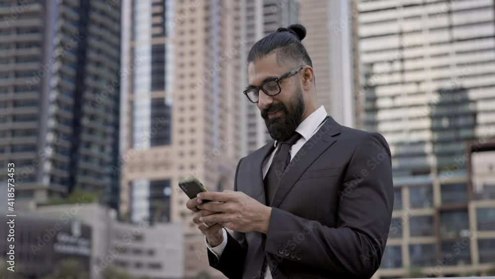Stylish Young Man In Business Suit On Background Of The City. Tall Buildings Dubai. He Holds Mobile Phone And Looks At It. Stylish Young Man In Business Suit On Background Of The City. Tall Buildings Dubai. He Holds Mobile Phone And Looks At It.