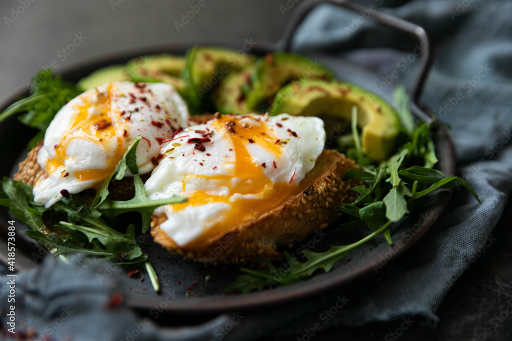 Delicious and delicious breakfast with poached egg on a slice of fried bread, avocado, arugula, cherry tomatoes and spices. Served in a cast-iron skillet, served with textiles on a black background . Delicious and delicious breakfast with poached egg on a slice of fried bread, avocado, arugula, cherry tomatoes and spices. Served in a cast-iron skillet, served with textiles on a black background .