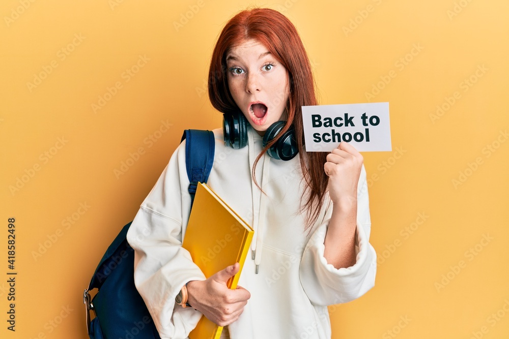 Young red head girl wearing backpack holding back to school banner afraid and shocked with surprise and amazed expression, fear and excited face. Young red head girl wearing backpack holding back to school banner afraid and shocked with surprise and amazed expression, fear and excited face.
