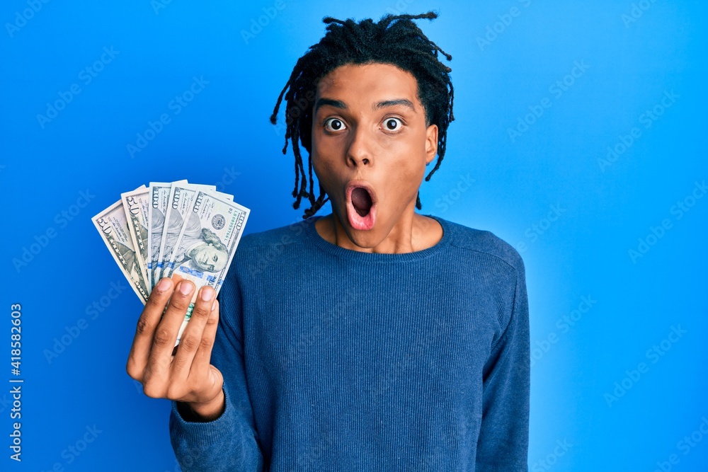Young african american man holding dollars scared and amazed with open mouth for surprise, disbelief face Young african american man holding dollars scared and amazed with open mouth for surprise, disbelief face