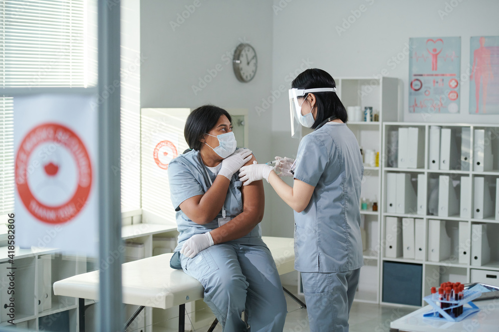 Young nurse in uniform and protective workwear making injection to her colleague Young nurse in uniform and protective workwear making injection to her colleague