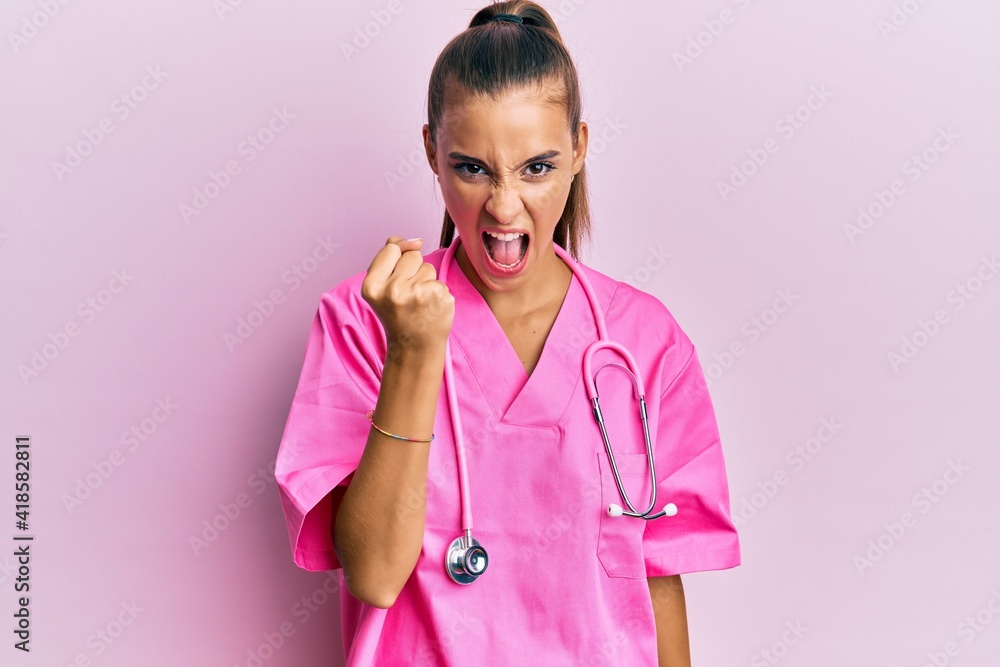 Young hispanic woman wearing doctor uniform and stethoscope angry and mad raising fist frustrated and furious while shouting with anger. rage and aggressive concept. Young hispanic woman wearing doctor uniform and stethoscope angry and mad raising fist frustrated and furious while shouting with anger. rage and aggressive concept.
