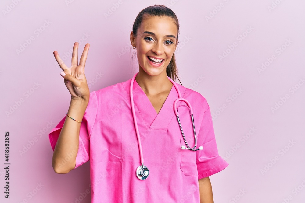 Young hispanic woman wearing doctor uniform and stethoscope showing and pointing up with fingers number three while smiling confident and happy. Young hispanic woman wearing doctor uniform and stethoscope showing and pointing up with fingers number three while smiling confident and happy.