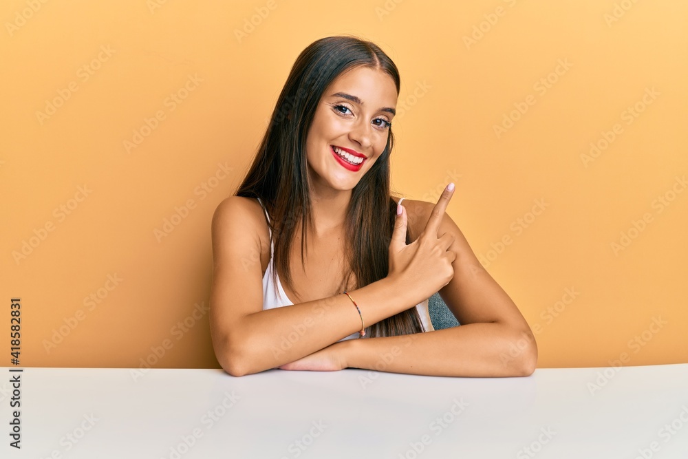 Young hispanic woman wearing casual clothes sitting on the table smiling cheerful pointing with hand and finger up to the side Young hispanic woman wearing casual clothes sitting on the table smiling cheerful pointing with hand and finger up to the side