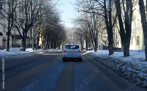 Spring sunny day. A white passenger car is driving along the road between the yellow houses. There is fresh snow on the roadsides.