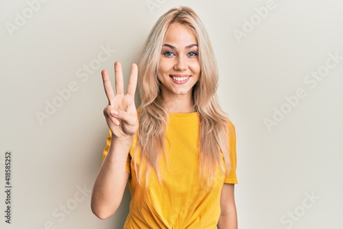Tela Beautiful caucasian blonde girl wearing casual tshirt showing and pointing up with fingers number three while smiling confident and happy