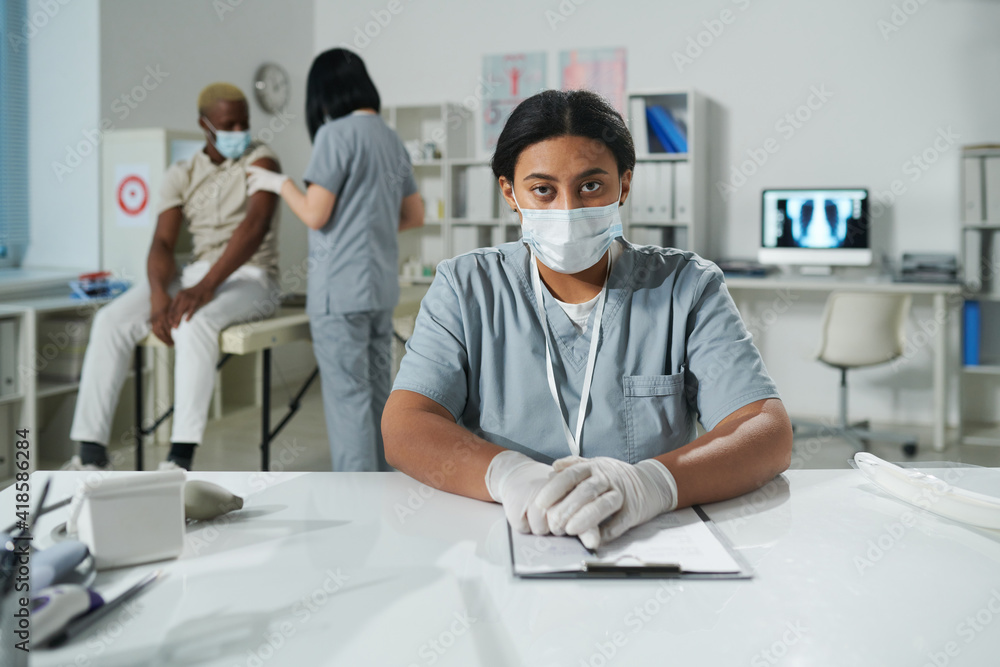 Young serious mixed-race female clinician in protective workwear sitting by desk Young serious mixed-race female clinician in protective workwear sitting by desk