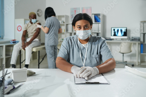Young serious mixed-race female clinician in protective workwear sitting by desk