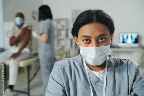 Young serious mixed-race female clinician in uniform and protective mask