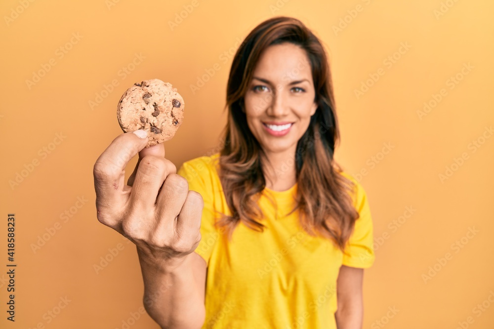Young latin woman holding cookie looking positive and happy standing and smiling with a confident smile showing teeth Young latin woman holding cookie looking positive and happy standing and smiling with a confident smile showing teeth