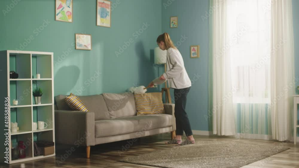 Pretty lady in a white sweater in a spacious bright room the color of a sea wave, with a window, wipes the dust from the lamp, beige sofa and white bookcase. Long shot Pretty lady in a white sweater in a spacious bright room the color of a sea wave, with a window, wipes the dust from the lamp, beige sofa and white bookcase. Long shot