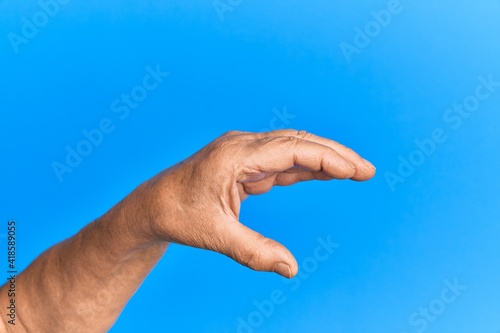 Hand of senior hispanic man over blue isolated background picking and taking invisible thing, holding object with fingers showing space
