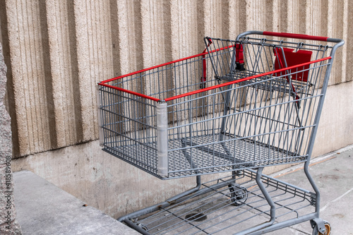 A silver and red metal shopping cart parked against a beige wall outside a supermarket in London, Ontario, Canada on a gloomy and overcast day winter day, 2021.
