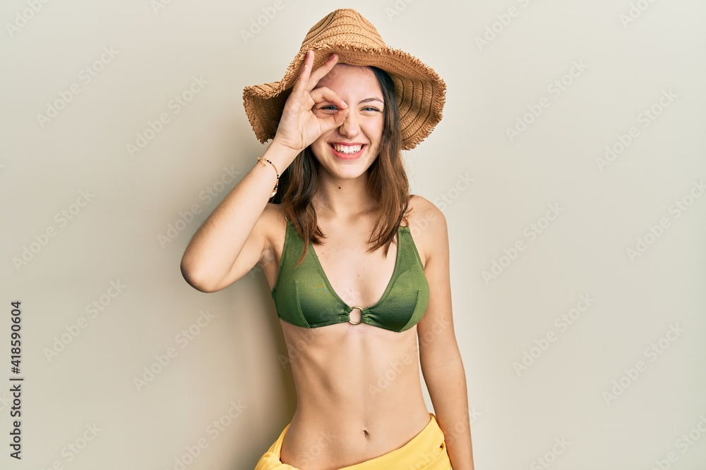 Young brunette woman wearing bikini doing ok gesture with hand smiling, eye looking through fingers with happy face. Young brunette woman wearing bikini doing ok gesture with hand smiling, eye looking through fingers with happy face.