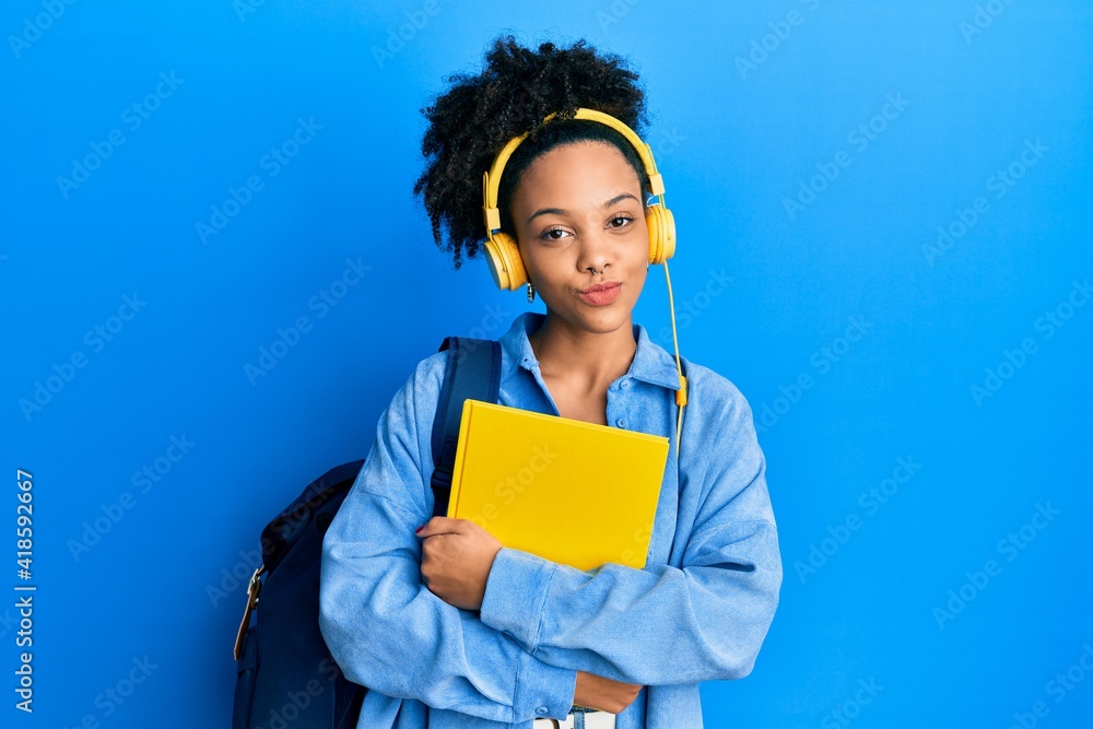 Obraz premium Young african american girl wearing student backpack and headphones holding book looking at the camera blowing a kiss being lovely and sexy. love expression.