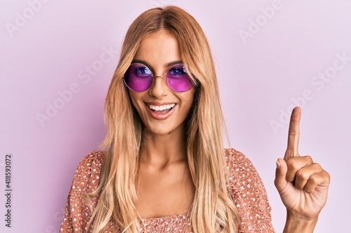Beautiful blonde young woman wearing fashion pink sunglasses smiling with an idea or question pointing finger with happy face, number one