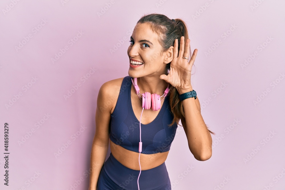 Young beautiful woman wearing gym clothes and using headphones smiling with hand over ear listening an hearing to rumor or gossip. deafness concept. Young beautiful woman wearing gym clothes and using headphones smiling with hand over ear listening an hearing to rumor or gossip. deafness concept.
