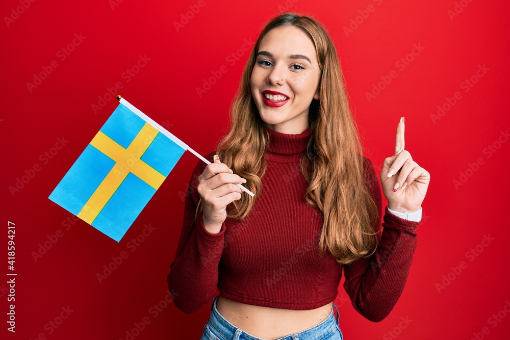 Young blonde woman holding sweden flag smiling with an idea or question pointing finger with happy face, number one Young blonde woman holding sweden flag smiling with an idea or question pointing finger with happy face, number one