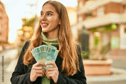 Young blonde girl smiling happy standing holding brazilian real banknotes at the city.