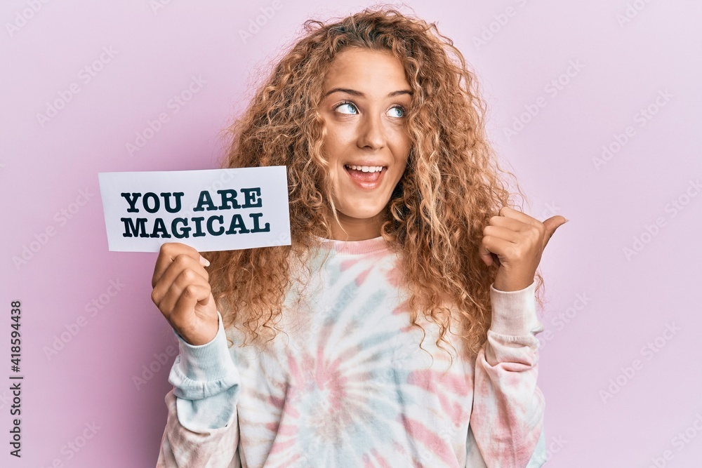 Beautiful caucasian teenager girl holding you are magical banner pointing thumb up to the side smiling happy with open mouth Beautiful caucasian teenager girl holding you are magical banner pointing thumb up to the side smiling happy with open mouth