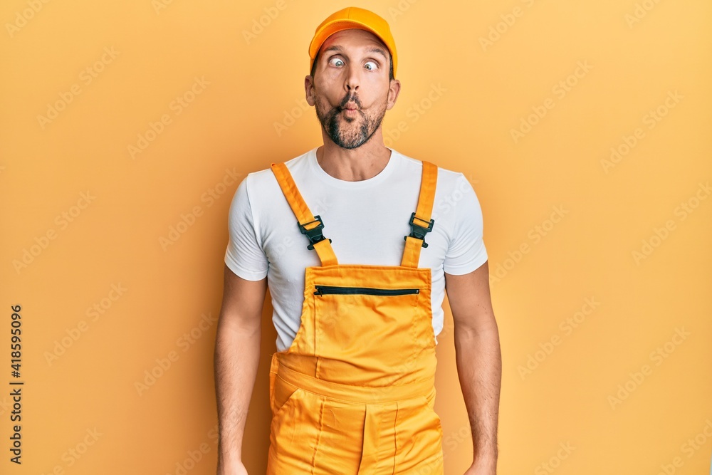 Young handsome man wearing handyman uniform over yellow background making fish face with lips, crazy and comical gesture. funny expression. Young handsome man wearing handyman uniform over yellow background making fish face with lips, crazy and comical gesture. funny expression.