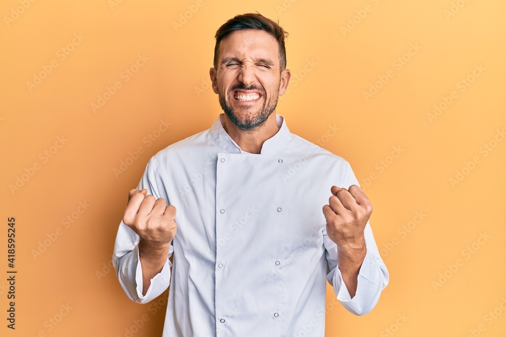 Handsome man with beard wearing professional cook uniform excited for success with arms raised and eyes closed celebrating victory smiling. winner concept. Handsome man with beard wearing professional cook uniform excited for success with arms raised and eyes closed celebrating victory smiling. winner concept.
