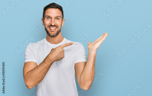 Handsome man with beard wearing casual white t shirt amazed and smiling to the camera while presenting with hand and pointing with finger.