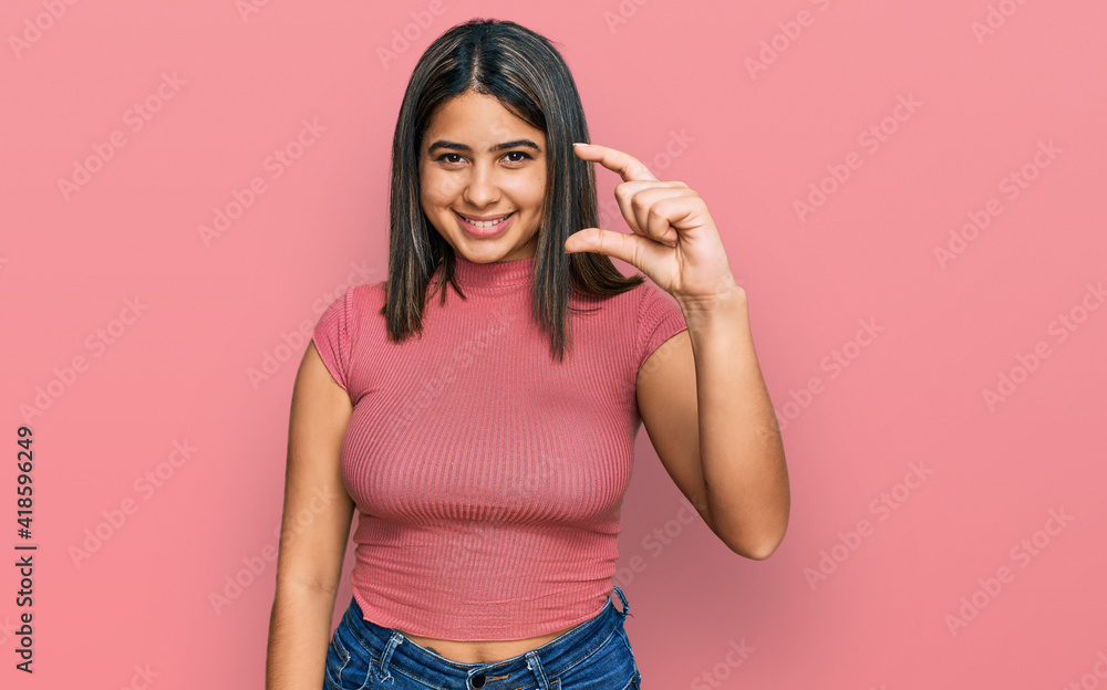 Young hispanic girl wearing casual t shirt smiling and confident gesturing with hand doing small size sign with fingers looking and the camera. measure concept. Young hispanic girl wearing casual t shirt smiling and confident gesturing with hand doing small size sign with fingers looking and the camera. measure concept.