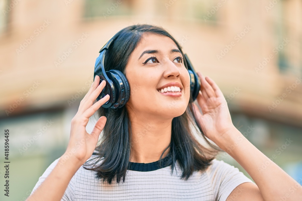 Young hispanic woman smiling happy listening to music using headphones at the city.