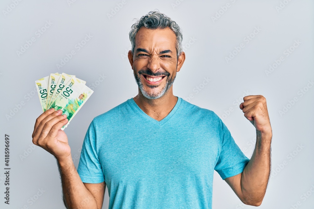 Middle age grey-haired man holding israel shekels banknotes screaming proud, celebrating victory and success very excited with raised arm Middle age grey-haired man holding israel shekels banknotes screaming proud, celebrating victory and success very excited with raised arm