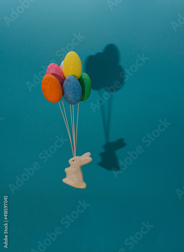 A white rabbit flies with balloons of colorful eggs. Blue background with shadow of bunny and easter eggs.